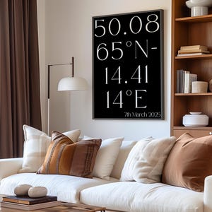 May include: A living room scene featuring a white sofa adorned with various throw pillows in shades of brown and cream. A wooden coffee table holds decorative items. A black framed print on the wall displays coordinates and the date 7th March 2025.