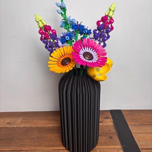 K&ouml;nnte beinhalten: Eine schwarze Vase mit vertikalen Rippen h&auml;lt einen bunten Strau&szlig; Lego-Blumen. Die Blumen umfassen gelbe Sonnenblumen, rosa Gerbera, lila Lupinen und blaue Vergissmeinnicht.