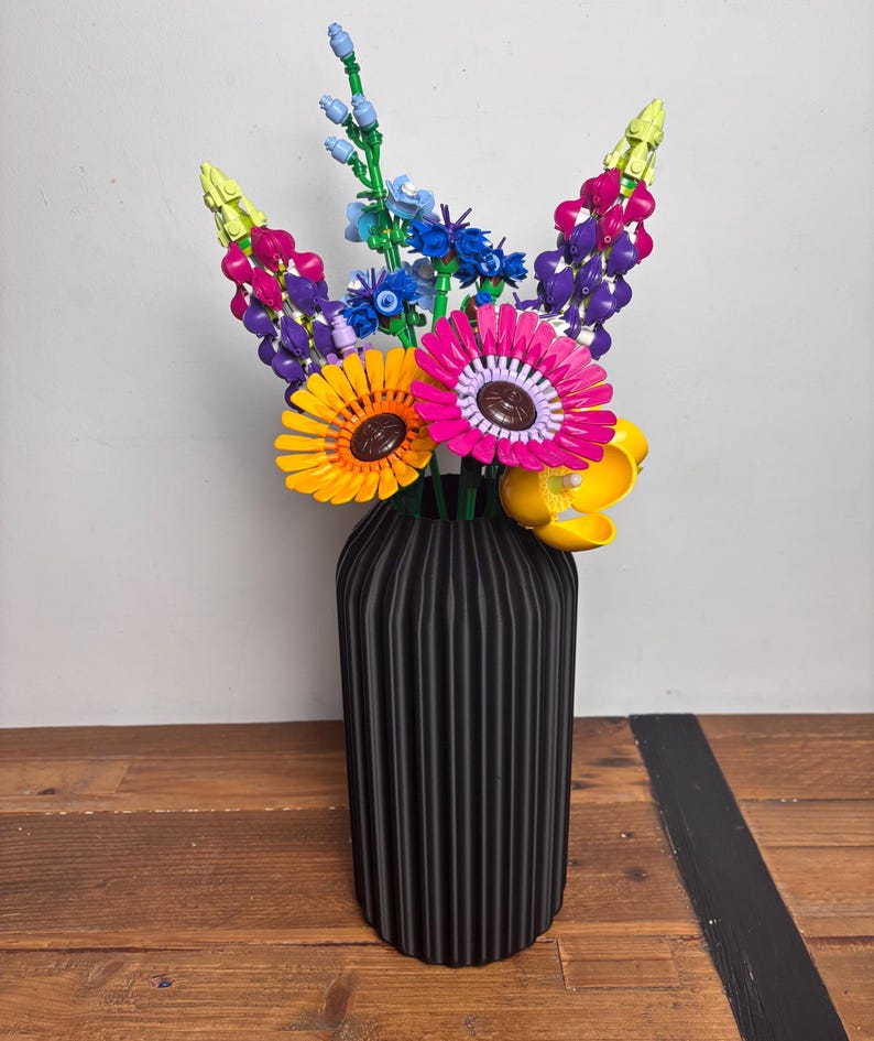 K&ouml;nnte beinhalten: Eine schwarze Vase mit vertikalen Rippen h&auml;lt einen bunten Strau&szlig; Lego-Blumen. Die Blumen umfassen gelbe Sonnenblumen, rosa Gerbera, lila Lupinen und blaue Vergissmeinnicht.