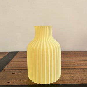 May include: A pale yellow ceramic vase with a fluted design. The vase is sitting on a wooden surface.