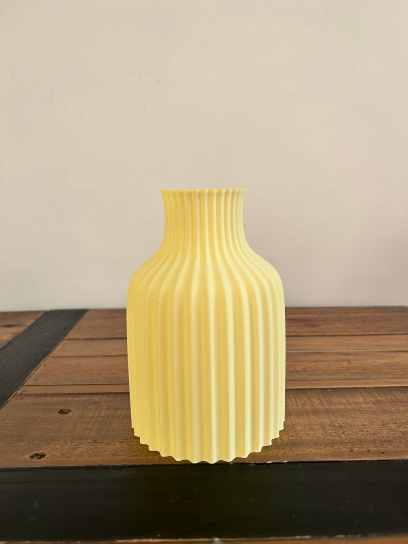May include: A pale yellow ceramic vase with a fluted design. The vase is sitting on a wooden surface.