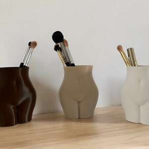 May include: Three ceramic vases shaped like a human torso, each in a different colour: brown, beige, and white. The vases are filled with makeup brushes.