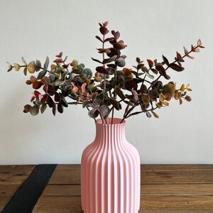 May include: A pink ceramic vase with a ribbed design, holding a bouquet of dried eucalyptus branches.