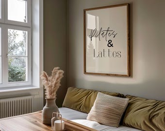 Puede incluir: Un cuadro enmarcado en madera con las palabras "Pilates & Lattes" en escritura negra. El cuadro cuelga de una pared de color neutro. Una mesa de madera en primer plano con un jarrón con flores secas y un candelabro.