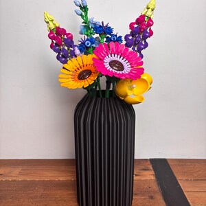 K&ouml;nnte beinhalten: Eine schwarze Vase mit vertikalen Rippen h&auml;lt einen Strau&szlig; bunter LEGO-Blumen. Die Blumen umfassen Gelb, Pink, Lila, Blau und Gr&uuml;n.