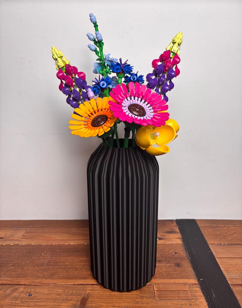 K&ouml;nnte beinhalten: Eine schwarze Vase mit vertikalen Rippen h&auml;lt einen Strau&szlig; bunter LEGO-Blumen. Die Blumen umfassen Gelb, Pink, Lila, Blau und Gr&uuml;n.