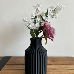 May include: A black vase with a ribbed design holds a bouquet of white flowers and a single pink rose.