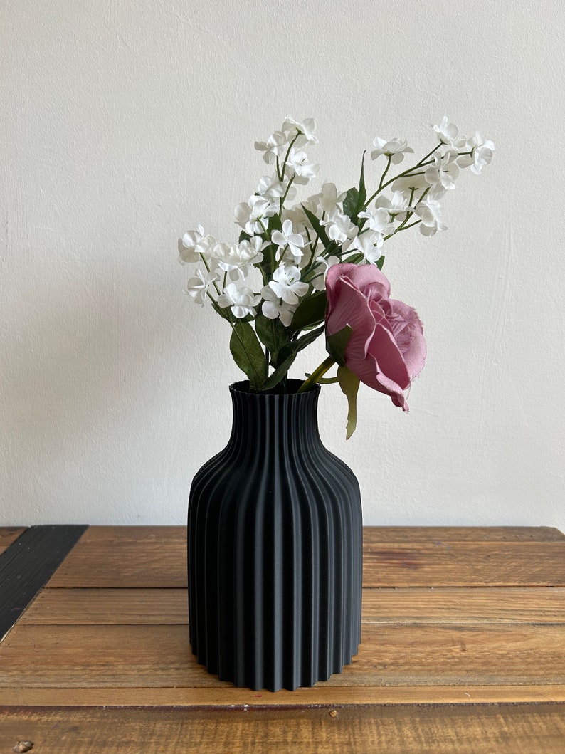 May include: A black vase with a ribbed design holds a bouquet of white flowers and a single pink rose.