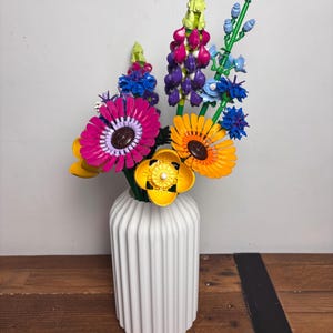 K&ouml;nnte beinhalten: Eine wei&szlig;e Keramikvase mit vertikalen Rippen h&auml;lt einen bunten Strau&szlig; Lego-Blumen. Die Blumen umfassen rosa, lila, gelbe, orangefarbene und blaue Bl&uuml;ten.