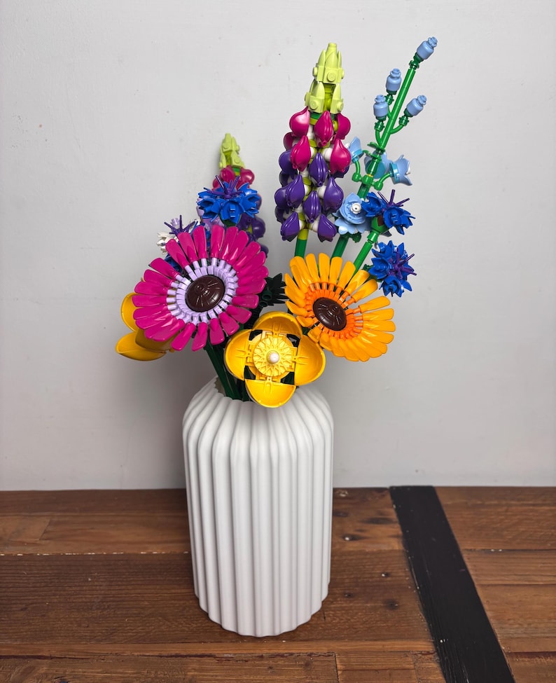 K&ouml;nnte beinhalten: Eine wei&szlig;e Keramikvase mit vertikalen Rippen h&auml;lt einen bunten Strau&szlig; Lego-Blumen. Die Blumen umfassen rosa, lila, gelbe, orangefarbene und blaue Bl&uuml;ten.