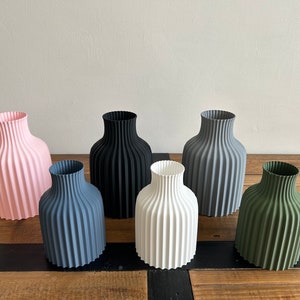 May include: Six ceramic vases in different colors, including pink, black, gray, white, and green. The vases have a ribbed design and are arranged in a row on a wooden surface.