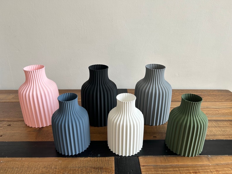 May include: Six ceramic vases in different colors, including pink, black, gray, white, and green. The vases have a ribbed design and are arranged in a row on a wooden surface.
