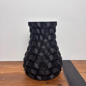 Op de afbeelding: Een zwarte keramische vaas met een geometrisch patroon. De vaas is gemaakt van een getextureerd materiaal en heeft een brede basis en een smalle hals.