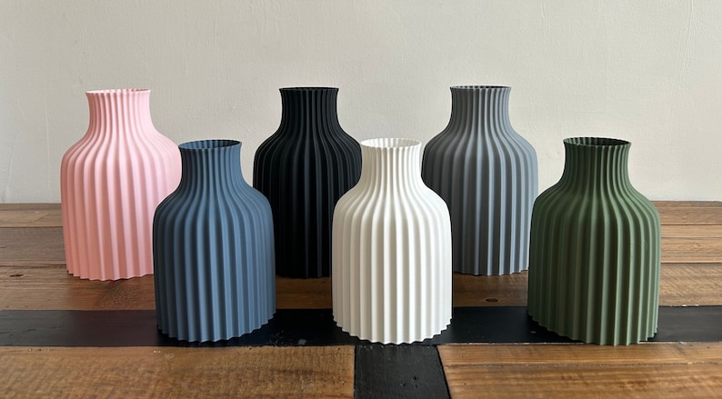 May include: Six ceramic vases in different colors, including pink, gray, black, white, and green. The vases have a ribbed design and are arranged in a row on a wooden surface.