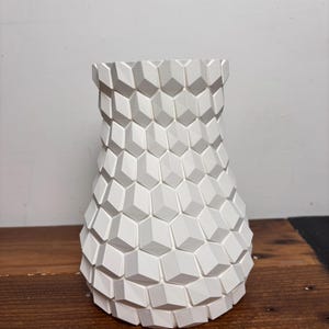 Op de afbeelding: Witte keramische vaas met een geometrisch, getextureerd ontwerp. De vaas heeft een brede basis en een smalle hals.
