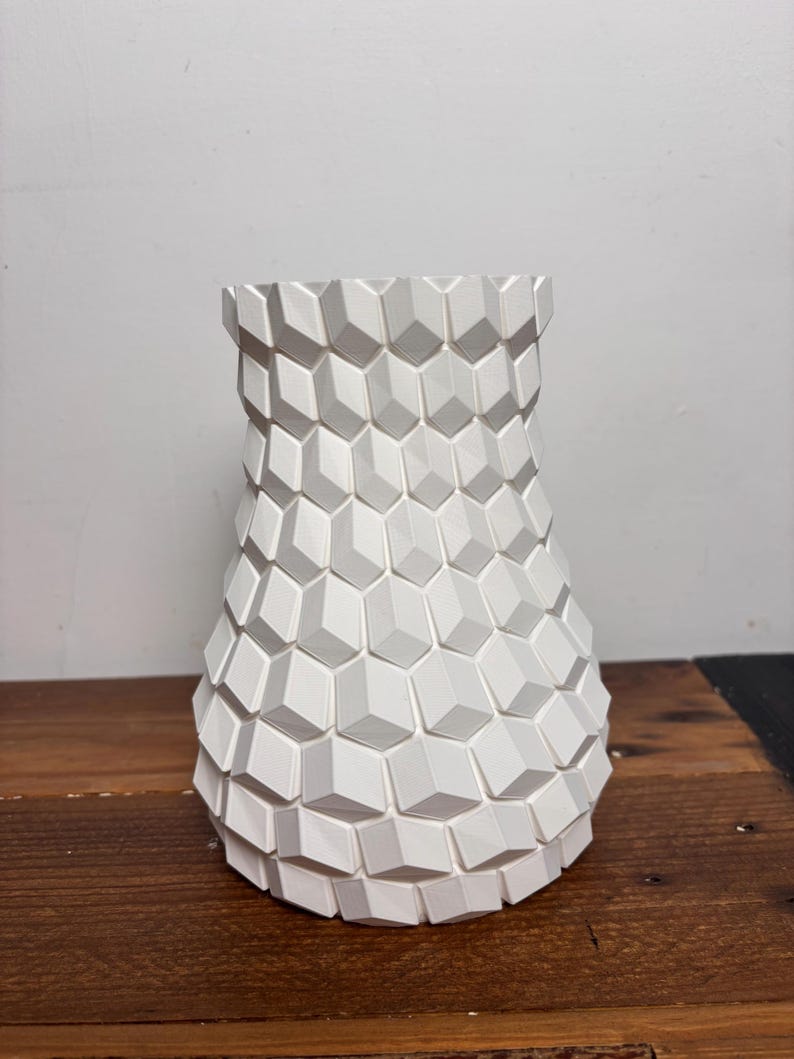 Op de afbeelding: Witte keramische vaas met een geometrisch, getextureerd ontwerp. De vaas heeft een brede basis en een smalle hals.