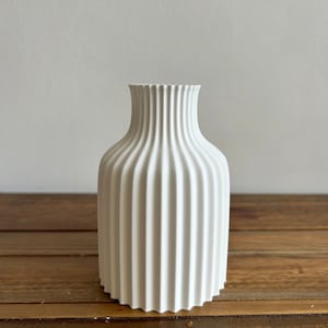 May include: A white ceramic vase with a fluted design. The vase is sitting on a wooden surface.