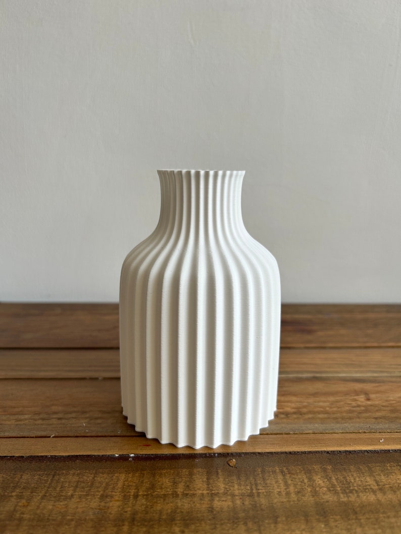 May include: A white ceramic vase with a fluted design. The vase is sitting on a wooden surface.