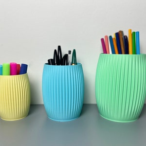 May include: Three cylindrical desk organisers in yellow, blue and green with a ribbed texture. The organisers are filled with pens, pencils and markers.