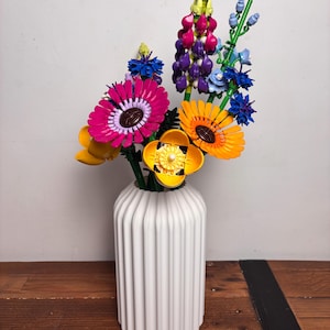 K&ouml;nnte beinhalten: Eine wei&szlig;e Keramikvase mit vertikalen Rippen h&auml;lt einen bunten Strau&szlig; Lego-Blumen. Der Strau&szlig; enth&auml;lt rosa, lila, gelbe und blaue Blumen.