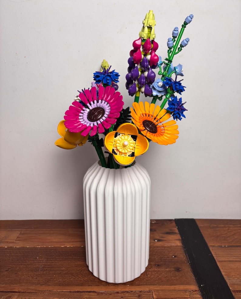 K&ouml;nnte beinhalten: Eine wei&szlig;e Keramikvase mit vertikalen Rippen h&auml;lt einen bunten Strau&szlig; Lego-Blumen. Der Strau&szlig; enth&auml;lt rosa, lila, gelbe und blaue Blumen.