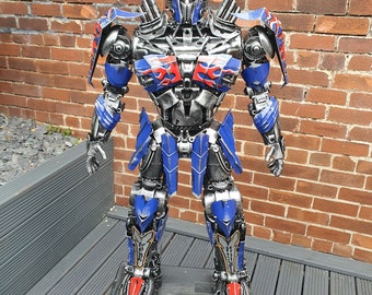 May include: A large, metallic sculpture of Optimus Prime, the iconic Transformers character. The sculpture features a detailed design with blue, silver, and red accents. It stands on a black base against a brick wall background.