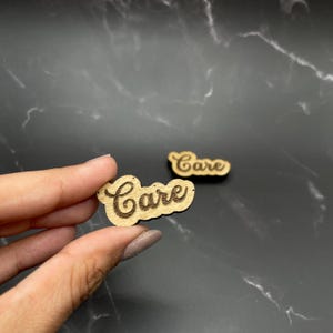 May include: Two small wooden cutouts with the word "Care" in a cursive font. One cutout is held by a hand, while the other lies on a dark, marbled surface. The wood has a light brown color with the word "Care" etched into the surface.