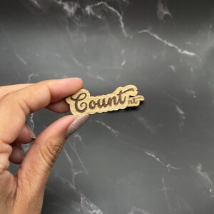 May include: A wooden cutout with the word "Count" written in a cursive font. The cutout is light brown and the lettering is dark brown.