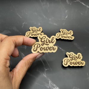 May include: Four wooden cutouts with the text "Girl Power" in a cursive font. The cutouts are brown and have a natural wood grain.
