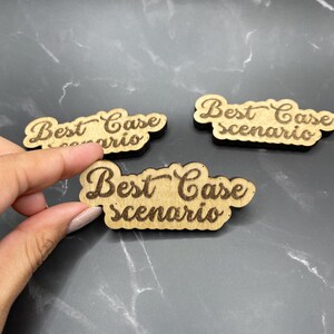 May include: Three wooden signs with the text "Best Case Scenario" in a cursive font. The signs are light brown and have a slightly rough texture.