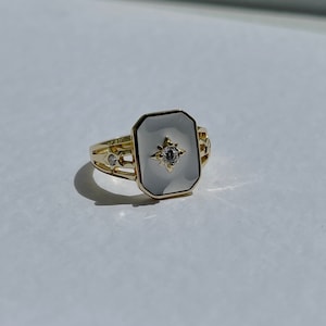 Könnte beinhalten: Ein goldener Ring mit einem rechteckigen weißen Stein, der in der Mitte eingesetzt ist. Der Stein hat einen kleinen diamantförmigen Stein in der Mitte. Der Ring hat ein dekoratives Design um den Stein herum.