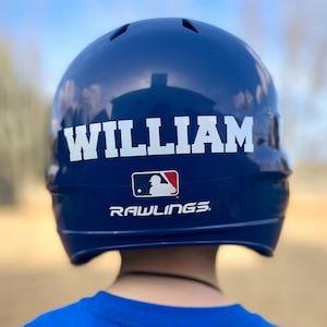 May include: A navy blue baseball helmet with the name "WILLIAM" in white block letters. The Rawlings logo and a Major League Baseball logo are also visible. The helmet is worn by a person wearing a blue shirt.