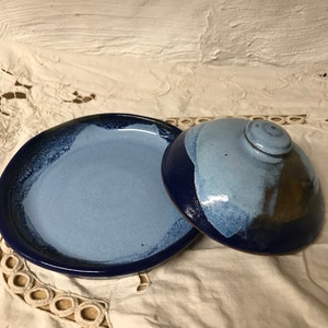 May include: A blue and white ceramic butter dish with a lid. The dish has a light blue glaze with a darker blue rim. The lid has a knob on top.