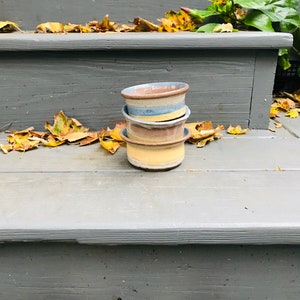 May include: Three stacked ceramic bowls with a brown, blue, and yellow striped design. The bowls are sitting on a gray wooden step with fallen autumn leaves.