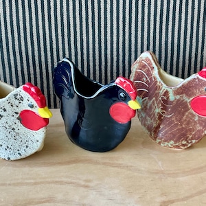 May include: Three ceramic chicken-shaped planters or containers. The first is white with black speckles, the second is solid glossy black, and the third is brown with a mottled finish. Each features a red comb, red wattle, and yellow beak.