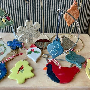 May include: A collection of ceramic Christmas ornaments in various shapes, including a snowflake, a tree with lights, and a cardinal bird. The ornaments are hand-painted with bright colors and festive designs, perfect for holiday decorating.