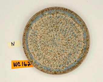 May include: A round ceramic plate with a textured surface. The plate features a speckled beige and brown center with a light blue rim. The rim and outer edge are a darker brown color. The plate has the text "WC 162" on a yellow sticker.