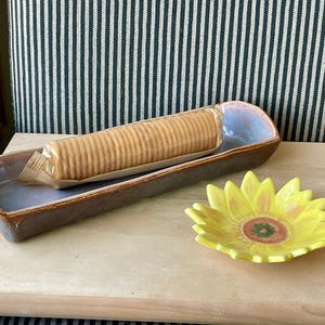 May include: A rectangular ceramic serving dish with a brown exterior and blue interior holds a package of crackers. A yellow sunflower-shaped dish sits beside it. The items are on a light-colored wooden surface.