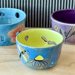 May include: Three ceramic bowls with different floral and insect designs. The bowls are blue, purple, and yellow with hand-painted details.