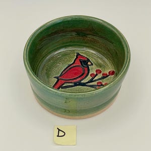 May include: A handmade ceramic bowl with a green exterior and interior. The interior features a red cardinal bird design with black accents and red berries. The bowl has a natural, unglazed base and a smooth, glossy finish.