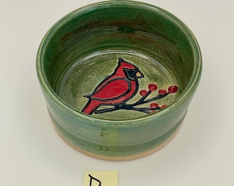 May include: A handmade ceramic bowl with a green exterior and interior. The interior features a red cardinal bird design with black accents and red berries. The bowl has a natural, unglazed base and a smooth, glossy finish.