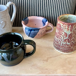 Puede incluir: Cuatro tazas de cerámica con diferentes diseños y colores. Una taza es blanca con un diseño de corazón, otra es rosa con diseños abstractos azules y blancos, otra es verde con letras negras que dicen "LOVE" y la última es marrón con un patrón detallado.