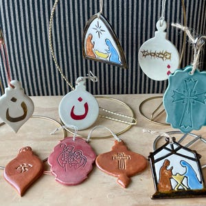 Pode incluir: Uma coleção de enfeites de Natal de cerâmica em várias formas e designs. Os enfeites apresentam símbolos religiosos, incluindo uma cruz, uma cena da natividade e caligrafia árabe. A paleta de cores inclui branco, vermelho, castanho e azul.