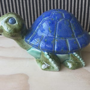 Puede incluir: Figura de tortuga de cerámica con caparazón azul brillante y cabeza, patas y cola verdes. La tortuga tiene un diseño de caparazón detallado y texturizado, colocada sobre una superficie clara.