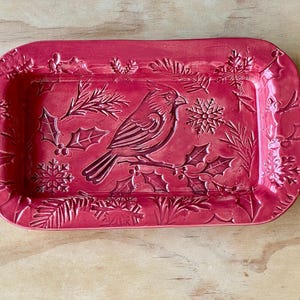 May include: A rectangular red ceramic tray featuring a raised, embossed design of a cardinal bird perched on a branch, surrounded by holly leaves, berries, and snowflakes. The tray has a glossy finish and rounded corners with a textured surface.