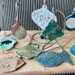 Peut inclure: Une collection de décorations de Noël en céramique de différentes formes, dont un poisson, un sapin et un bison. Les ornements sont dans des tons de bleu, vert et marron, avec des motifs décoratifs. Ils sont exposés sur une surface en bois.