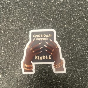 Pode incluir: Um autocolante com m&atilde;os de pele escura adornadas com an&eacute;is e pulseiras douradas segurando um ret&acirc;ngulo preto. O ret&acirc;ngulo diz "EMOTIONAL SUPPORT KINDLE" em texto branco. O autocolante est&aacute; sobre uma superf&iacute;cie cinza salpicada.