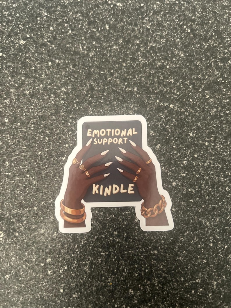 Pode incluir: Um autocolante com m&atilde;os de pele escura adornadas com an&eacute;is e pulseiras douradas segurando um ret&acirc;ngulo preto. O ret&acirc;ngulo diz "EMOTIONAL SUPPORT KINDLE" em texto branco. O autocolante est&aacute; sobre uma superf&iacute;cie cinza salpicada.