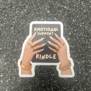 Pode incluir: Um autocolante com um ret&acirc;ngulo preto com as palavras "EMOTIONAL SUPPORT KINDLE" em branco. A imagem mostra m&atilde;os com unhas longas e brancas e joias douradas segurando o ret&acirc;ngulo.