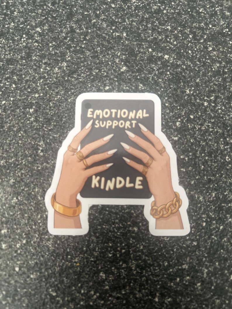 Pode incluir: Um autocolante com um ret&acirc;ngulo preto com as palavras "EMOTIONAL SUPPORT KINDLE" em branco. A imagem mostra m&atilde;os com unhas longas e brancas e joias douradas segurando o ret&acirc;ngulo.